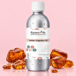A 100 mL bottle of Raasa Oils Amber Fragrance Oil, featuring a metallic silver body with water droplets, a white cap, and a white label with pink and green accents. The bottle is styled with several amber-colored crystals on a solid light pink background.