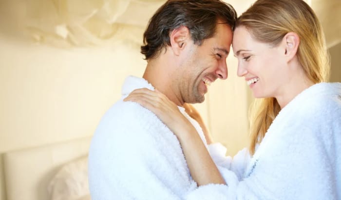 Couple in bathrobes smiling at each other in a bedroom.