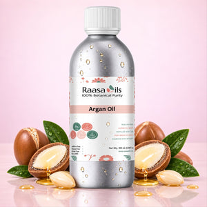 A bottle of Raasa Oils Argan Oil centered against a soft pink background, surrounded by whole and sliced argan nuts with visible golden oil and a few green leaves.