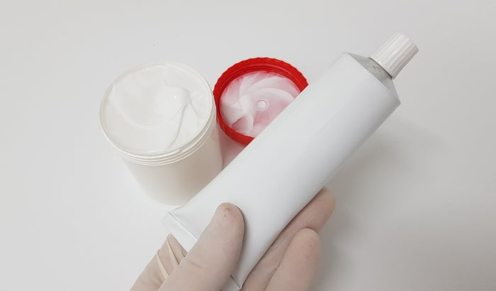 White bottle with a red cap and pink foam, held by a hand wearing a beige glove on a white background.