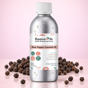 A bottle of Raasa oils Black Pepper Essential Oil centered on a light pink background, surrounded by scattered whole black peppercorns. The bottle is metallic silver with a white cap and a white label featuring pink and green accents.