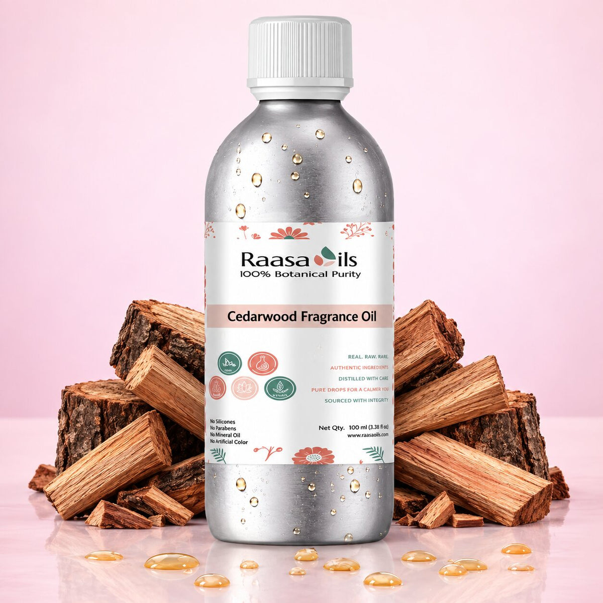 A bottle of Raasa oils Cedarwood Fragrance Oil stands centered on a pink background, flanked by stacked wood chunks and scattered droplets of the oil. The bottle is metallic silver with a white cap and a white label featuring botanical illustrations and product details.