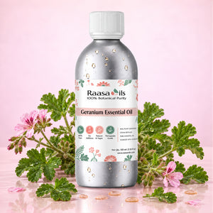 A bottle of Raasa Oils Geranium Essential Oil centered on a pink reflective surface, surrounded by green geranium leaves and pink flowers. The bottle is metallic silver with a white cap and a floral label.