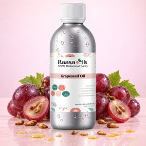 A bottle of Raasa oils Grapeseed Oil centered on a pink background, flanked by fresh red grapes, sliced grapes, and scattered oil droplets. The bottle is metallic silver with a white cap and a white label featuring pink and green accents.