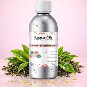 A bottle of Raasa Oils Green Tea Essential Oil centered on a pink background, surrounded by green tea leaves and loose tea buds with water droplets. The bottle is metallic silver with a white cap and a white label featuring green and pink accents.