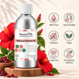 A bottle of Raasa Oils Hibiscus Essential Oil shown on a wooden stand with red hibiscus flowers around it. The bottle is metallic silver with a white cap and a white label featuring pink and green accents.