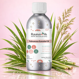 A bottle of Raasails Palmarosa Essential Oil centered on a pink background, surrounded by green leaves and dried palm fronds with water droplets. The bottle is metallic silver with a white cap and a white label featuring pink and green botanical details.