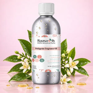 A bottle of Raasa Oils Petitgrain Fragrance Oil stands centered on a pink background, surrounded by green leaves, white flowers, and scattered golden oil droplets. The bottle is metallic silver with a white cap and a white label featuring pink floral accents and product details.