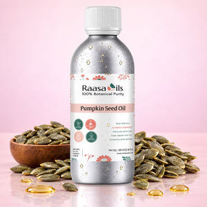 A bottle of Raasa Oils Pumpkin Seed Oil centered on a pink background, surrounded by scattered pumpkin seeds and a wooden bowl of seeds. The bottle is metallic silver with a white cap and a white label featuring pink floral accents and product text.