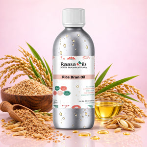A bottle of Raasa oils Rice Bran Oil centered against a soft pink background, surrounded by rice grains, a wooden bowl, a small glass of golden oil, and scattered oil droplets. The bottle is metallic silver with a white cap and a white label featuring the Raasa oils brand.