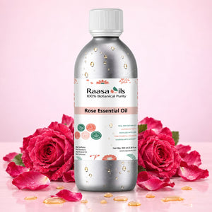 A bottle of Raasa oils Rose Essential Oil surrounded by fresh pink roses and scattered petals on a pink background.