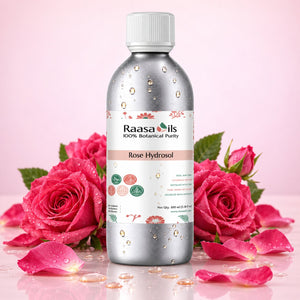 A bottle of Raasa Oils Rose Hydrosol centered on a pink background, surrounded by fresh pink roses, petals, and water droplets. The bottle is metallic silver with a white cap and a white label featuring pink floral accents.
