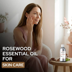Woman applying rosewood essential oil to her face with a bottle of the product on a table.