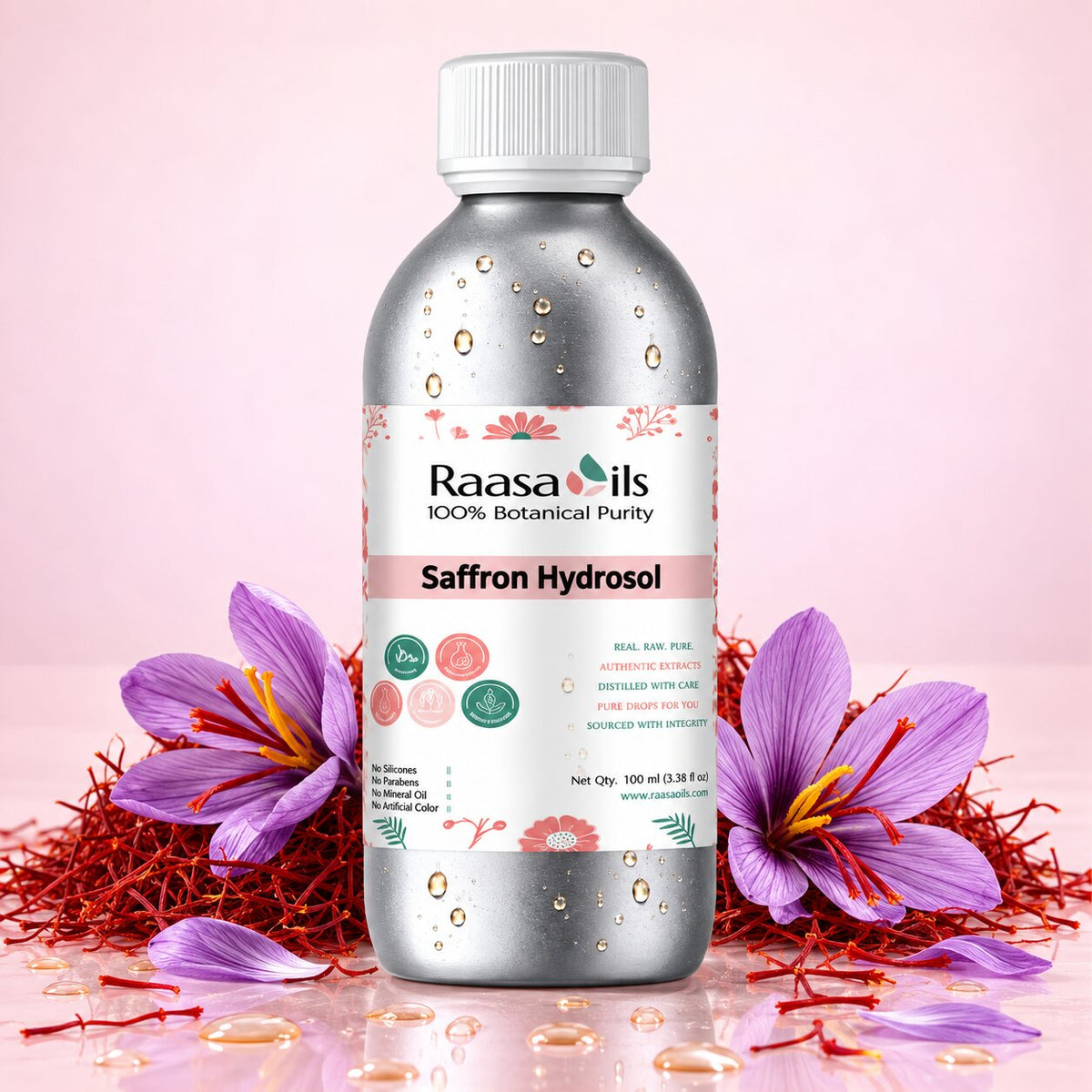 A 100 ml bottle of Raasa Oils Saffron Hydrosol, featuring a metallic silver bottle with a white cap and a white label decorated with pink saffron flowers. The bottle is styled with purple saffron flowers and red saffron threads on a soft pink background.