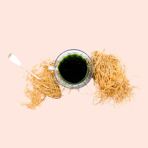 A small glass bottle filled with a green liquid, surrounded by dry, straw-like roots, on a light background.