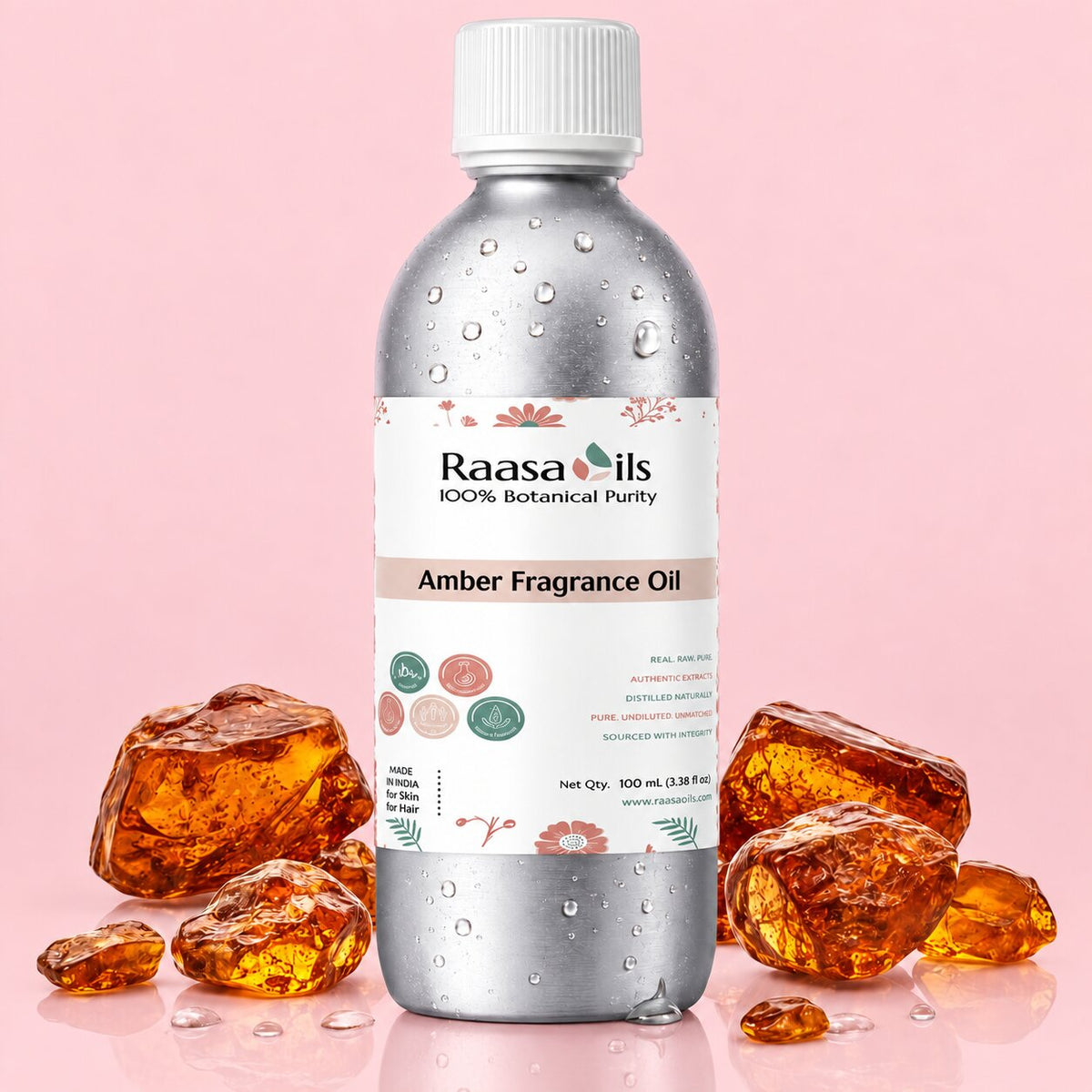A 100 mL bottle of Raasa Oils Amber Fragrance Oil, featuring a metallic silver body with water droplets, a white cap, and a white label with pink and green accents. The bottle is styled with several amber-colored crystals on a solid light pink background.