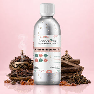 A bottle of Raasa oils Bakhoor Fragrance Oil centered on a pink background, surrounded by decorative incense, wood pieces, and amber resin pieces. The bottle is metallic silver with a white cap and a white label featuring pink and green botanical illustrations.
