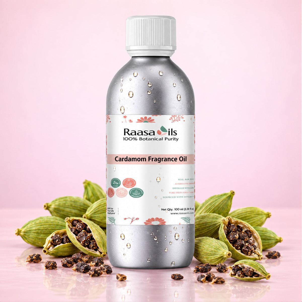 A bottle of Raasails Cardamom Fragrance Oil centered on a pink background, surrounded by whole and halved green cardamom pods with dark seeds. The bottle is metallic silver with a white cap and a white label featuring pink floral accents and product text.