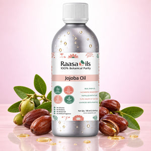 A bottle of Raasa Oils Jojoba Oil centered on a soft pink background, surrounded by whole and cracked jojoba seeds with green leaves. The bottle is metallic silver with a white cap and a white label featuring pink and green accents.