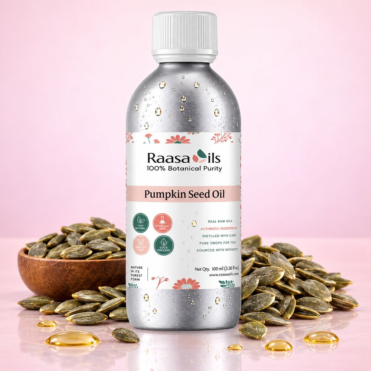 A bottle of Raasa Oils Pumpkin Seed Oil centered on a pink background, surrounded by scattered pumpkin seeds and a wooden bowl of seeds. The bottle is metallic silver with a white cap and a white label featuring pink floral accents and product text.