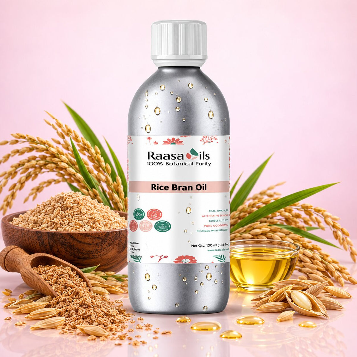 A bottle of Raasa oils Rice Bran Oil centered against a soft pink background, surrounded by rice grains, a wooden bowl, a small glass of golden oil, and scattered oil droplets. The bottle is metallic silver with a white cap and a white label featuring the Raasa oils brand.