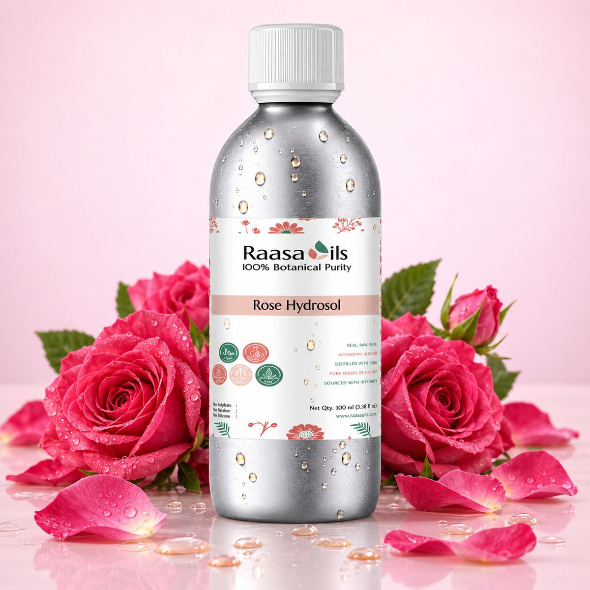 A bottle of Raasa Oils Rose Hydrosol centered on a pink background, surrounded by fresh pink roses, petals, and water droplets. The bottle is metallic silver with a white cap and a white label featuring pink floral accents.